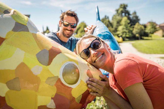 Ein Mann und eine Frau stehen vor einer bunt bemalten Fisch-Skulptur. Die Frau umarmt den Fisch. 