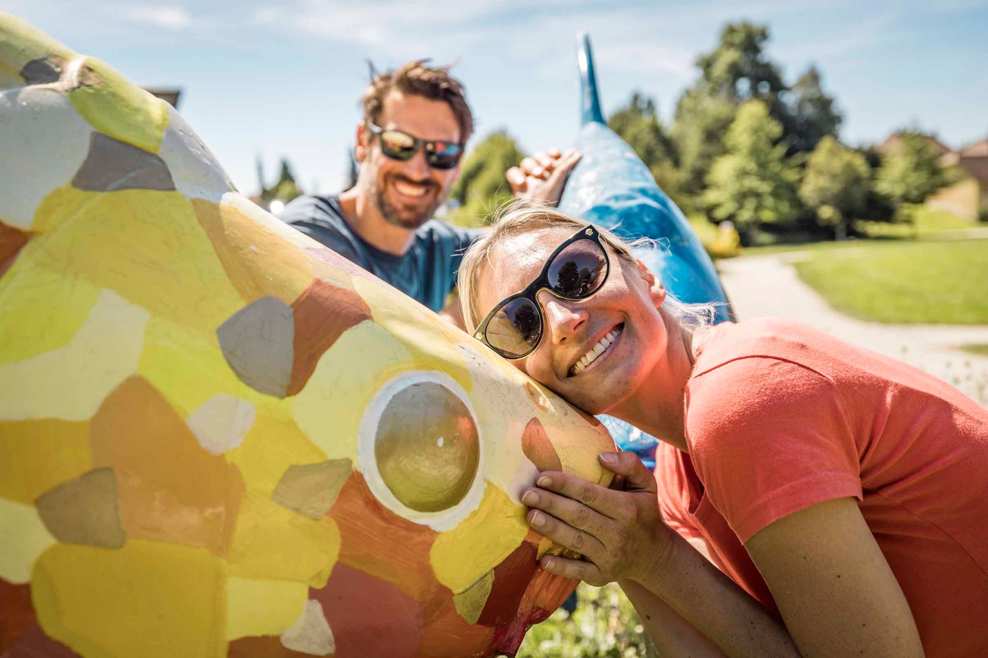 Ein Mann und eine Frau stehen vor einer bunt bemalten Fisch-Skulptur. Die Frau umarmt den Fisch. 