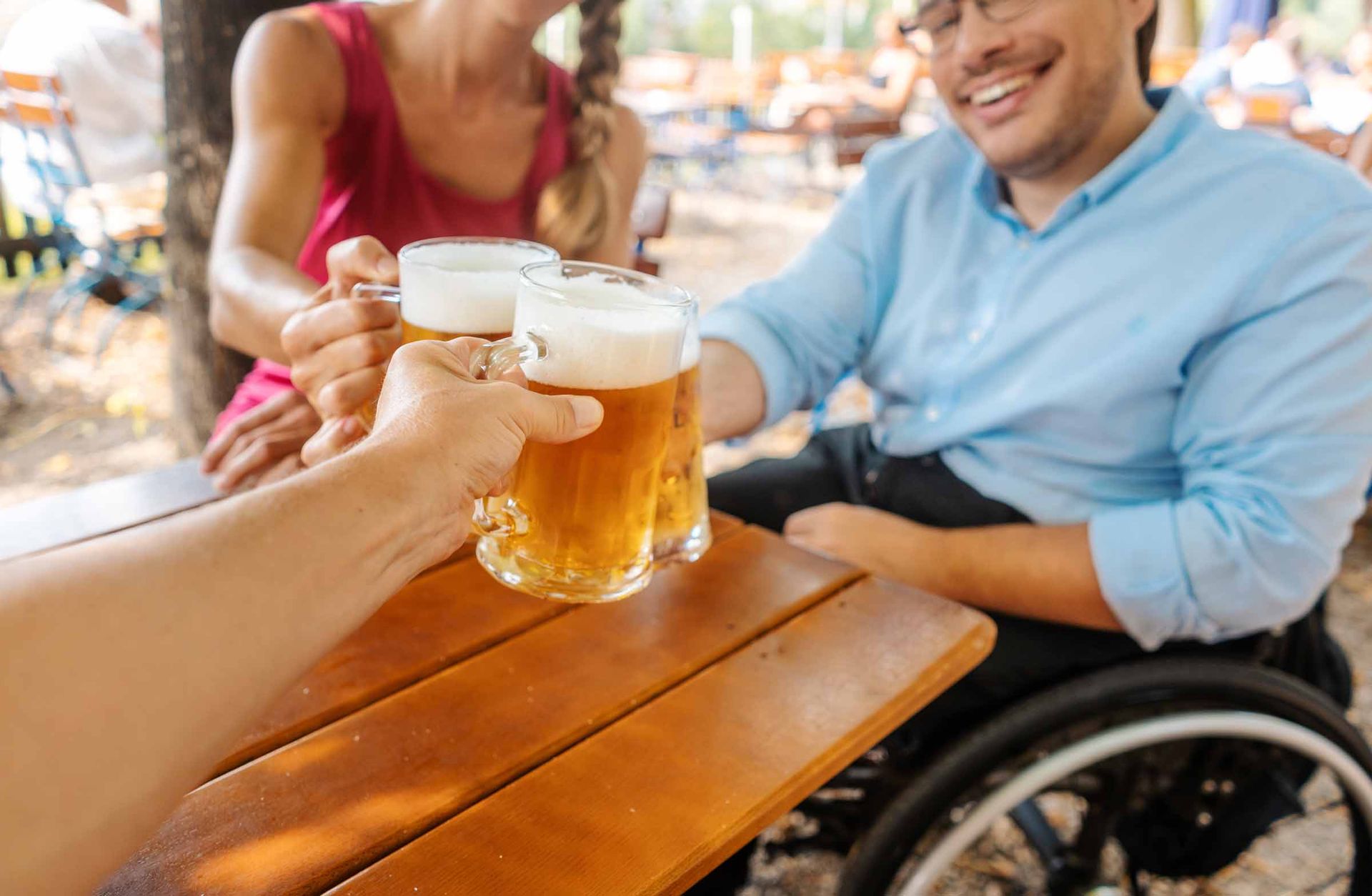 Drei Menschen stoßen im Biergarten mit Bierkrügen an. Eine Person, ein Mann, sitzt im Rollstuhl. Eine Person ist eine Frau. Von der dritten Person ist nur der Arm zu sehen. 
